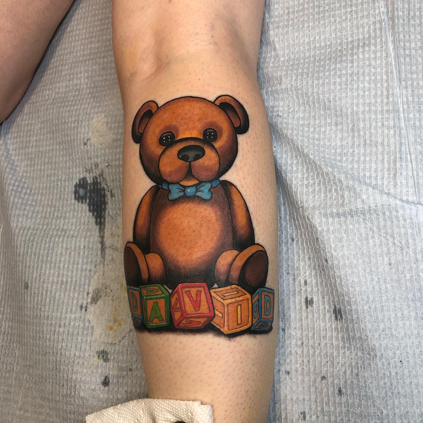 Tattoo of a teddy bear with a blue bowtie sitting atop colorful blocks spelling "DAVID"